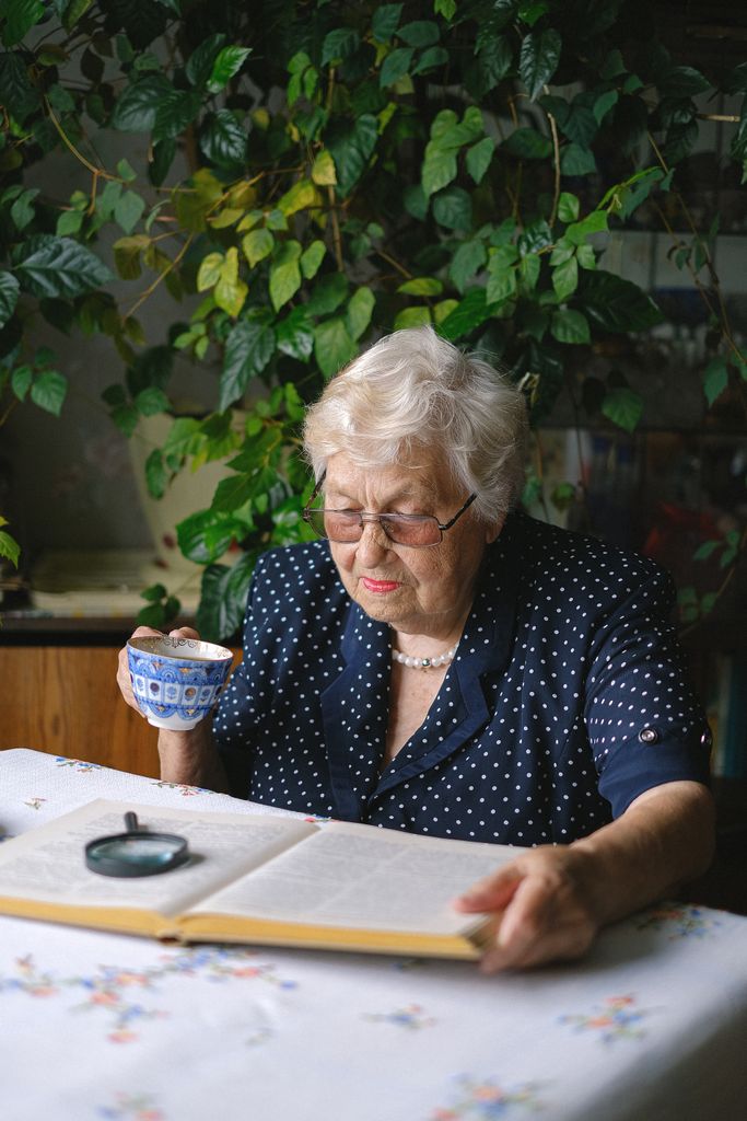 Accueil Focused aged female in eyeglasses sitting at table with book and magnifier while drinking tea in room with green plants